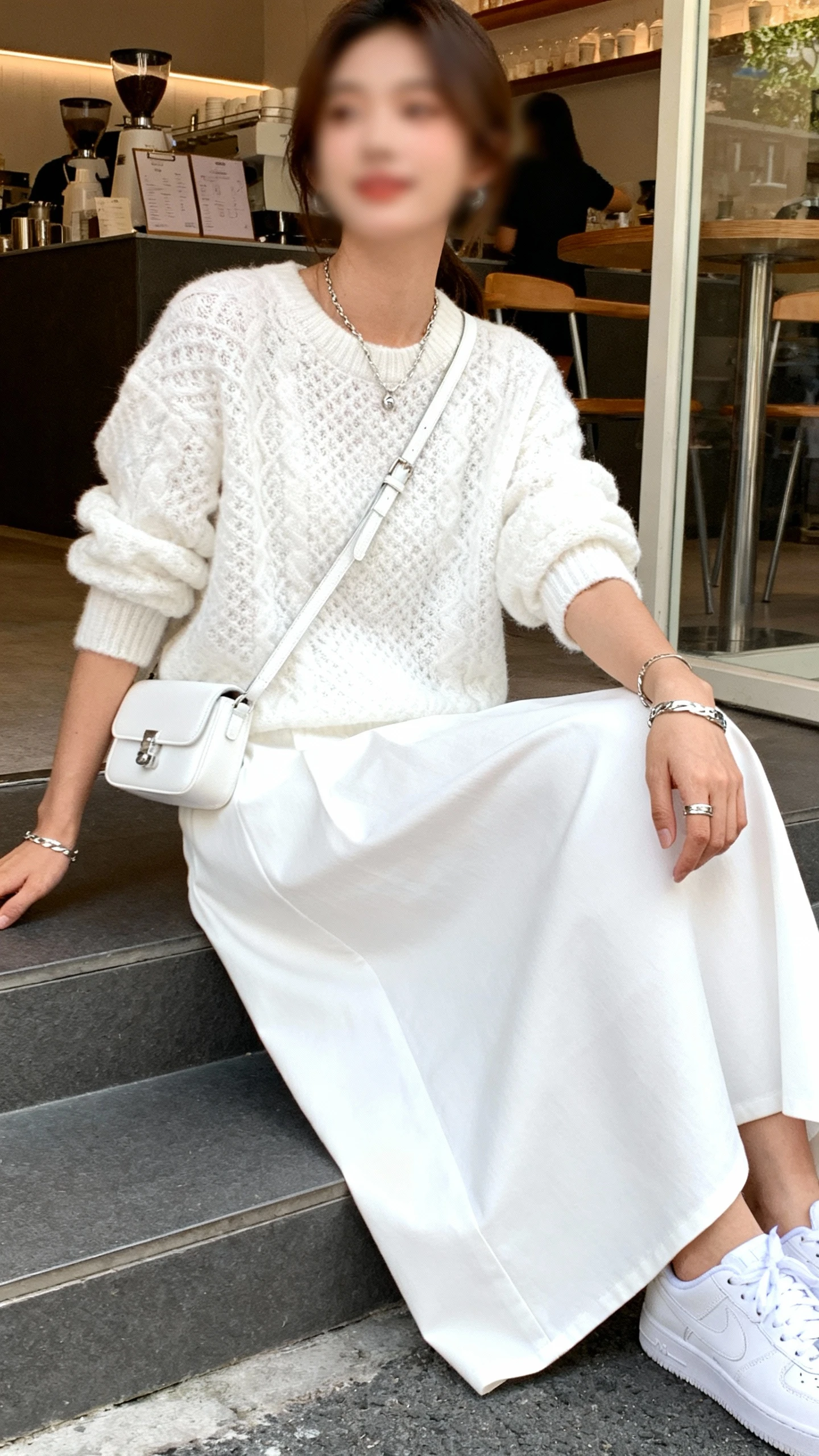 Candid photo of a woman wearing a monochrome white outfit: white maxi skirt with a textured white sweater and white sneakers, white crossbody and subtle silver jewelry, sitting on cafe steps, face slightly blurred, natural daylight, iPhone photo quality.