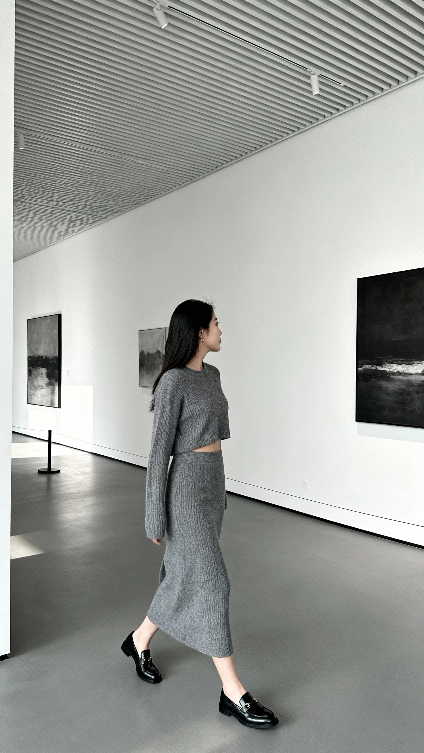 Minimalist lifestyle photo of a woman wearing a monochrome grey knit top and matching grey skirt set with sleek black loafers, walking through a modern gallery, face looking away, ambient daylight, iPhone photo quality.