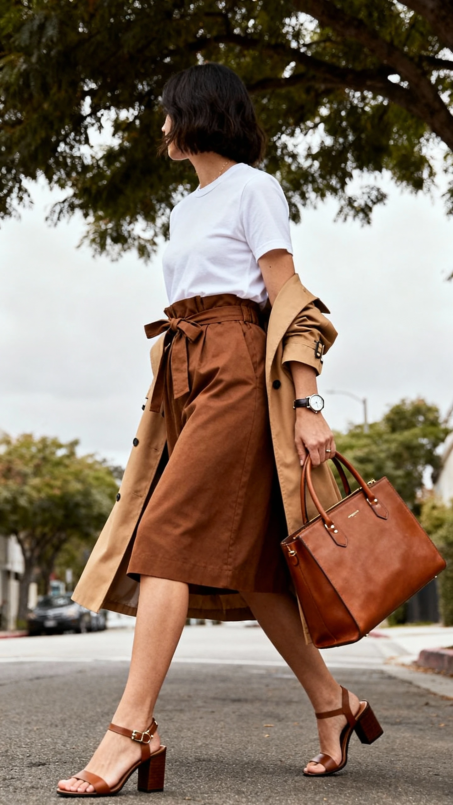 Natural lifestyle photo of a woman in a brown belted paperbag skirt, crisp white crewneck tee, block-heel sandals, classic tan trench, leather top-handle bag, simple watch and studs, walking under tree-lined street, face looking away, overcast daylight, iPhone photo quality.