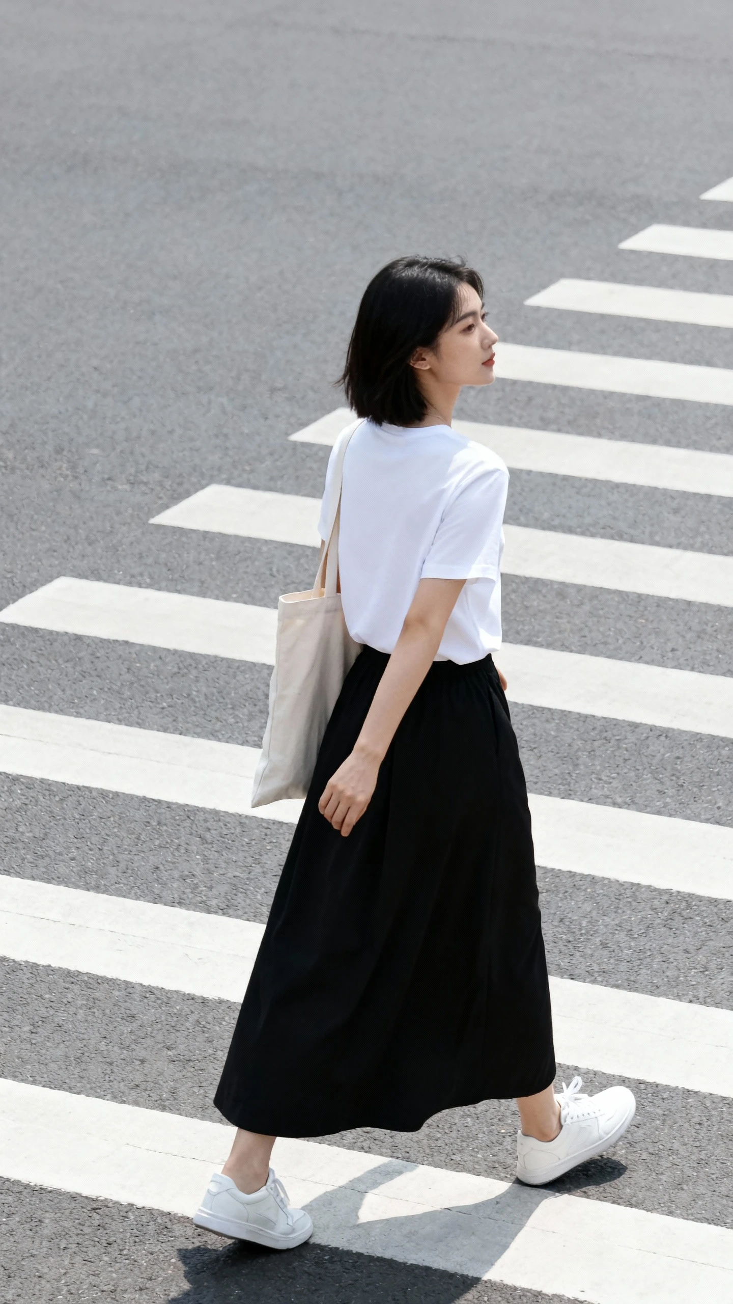 Natural lifestyle photo of a woman wearing a black maxi skirt with a crisp white tee and white sneakers, walking across a crosswalk with a tote bag, face looking away, natural daylight, iPhone photo quality, candid and unstaged.
