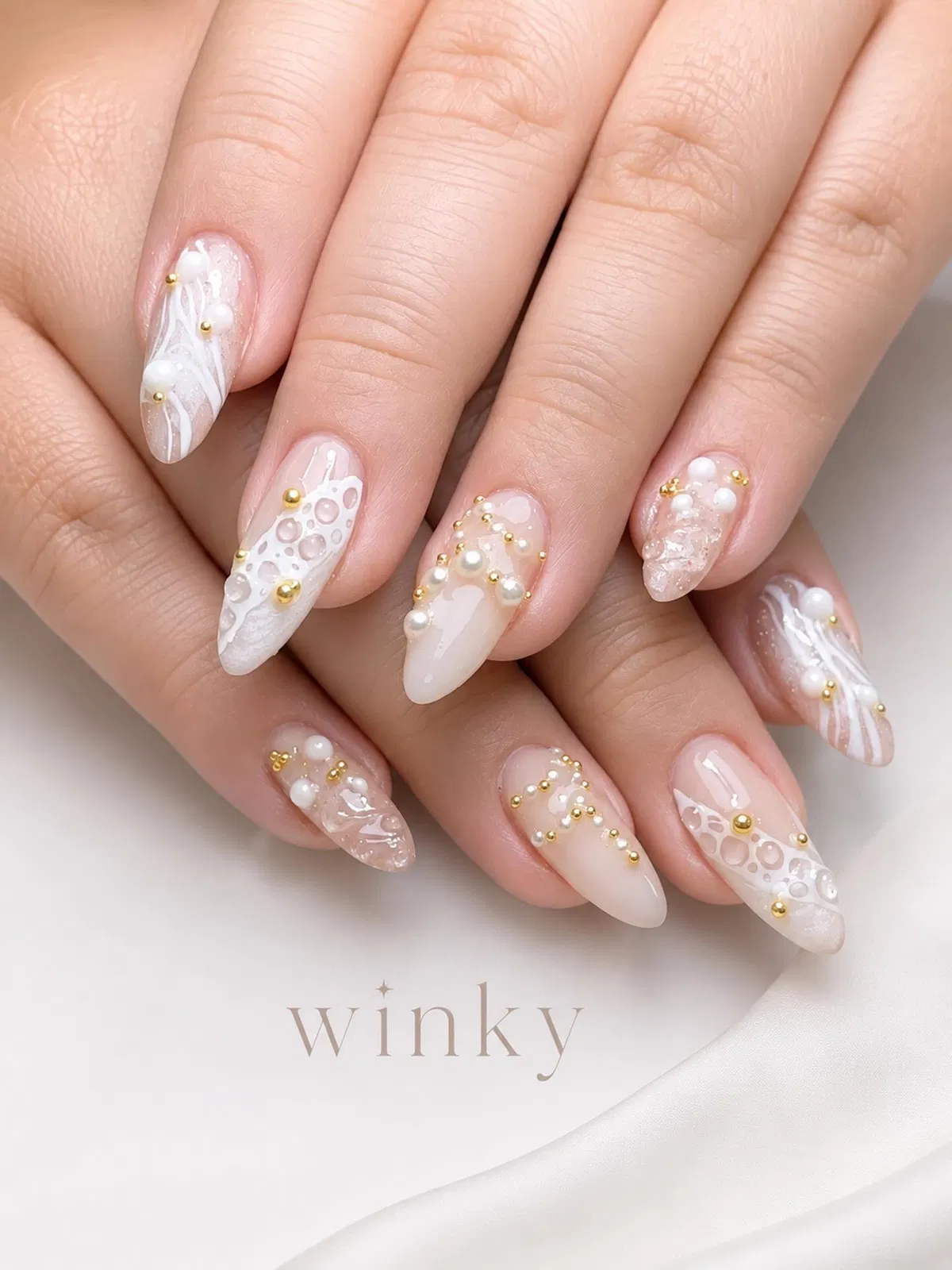 Sheer nude-pink almond nails with white swirls, pearls, and gold beads