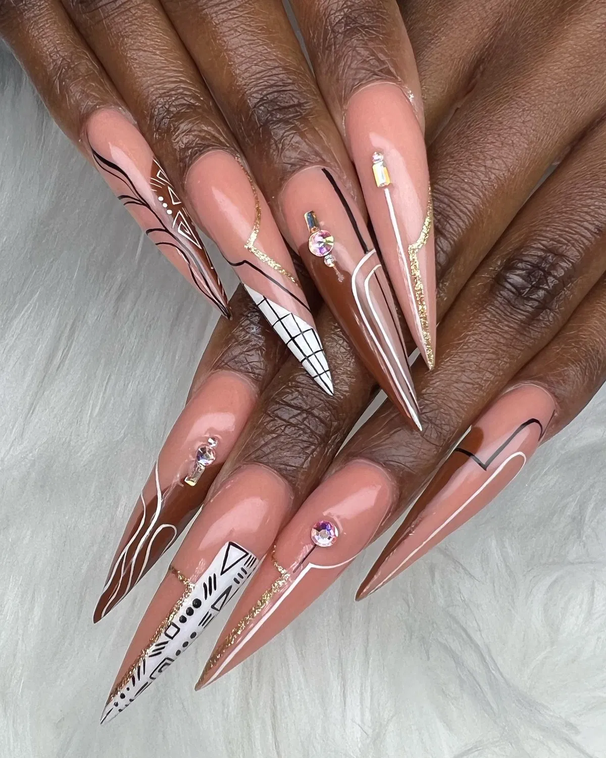Extra-long stiletto nails with nude-and-brown graphics, gold glitter edging, and rhinestones