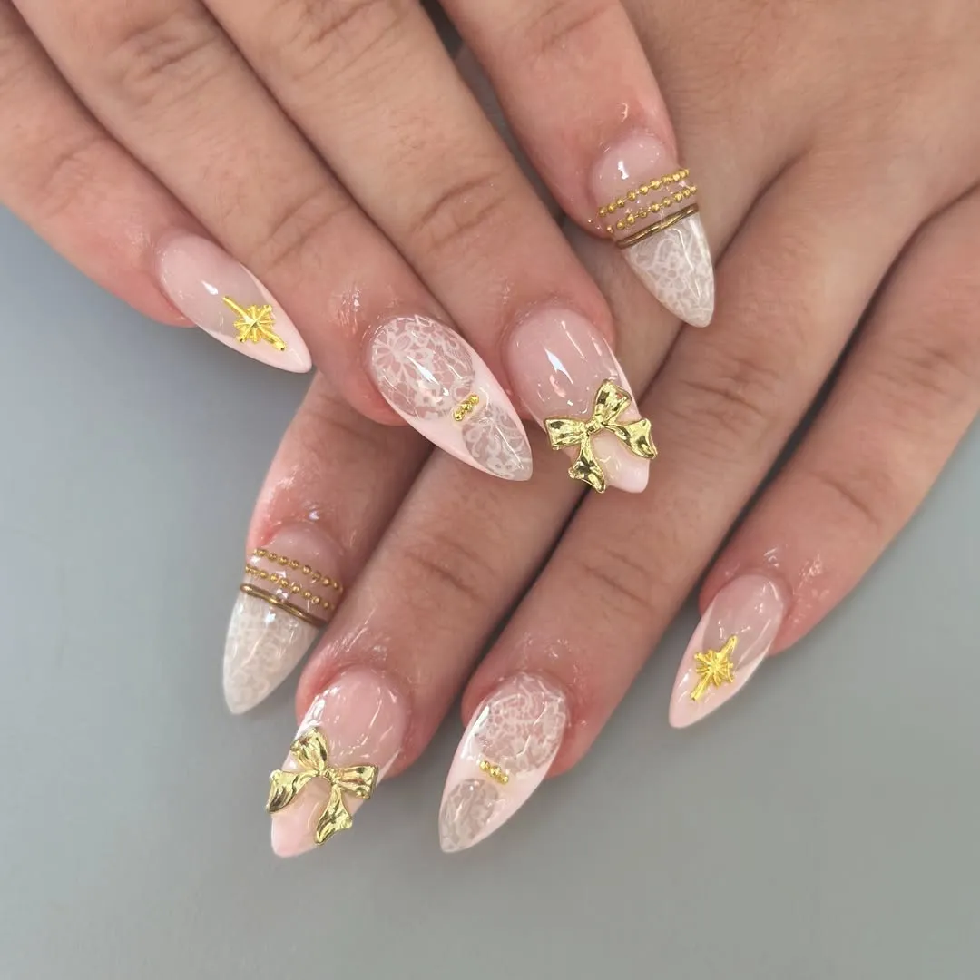 Pale pink almond nails with lace-like detail and gold charms
