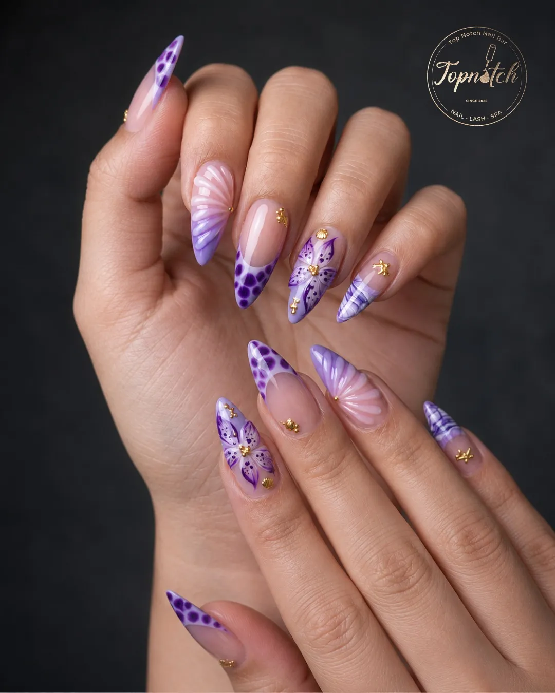 Purple patterned stiletto tips with small gold charms (logo overlay)