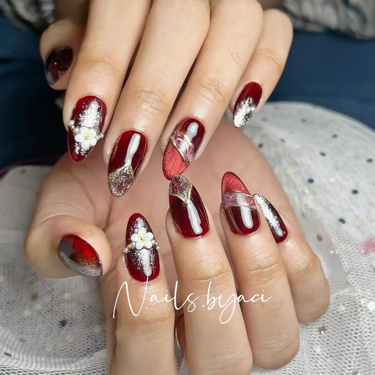 Almond maroon nails with silver glitter, gold lines, and 3D flowers