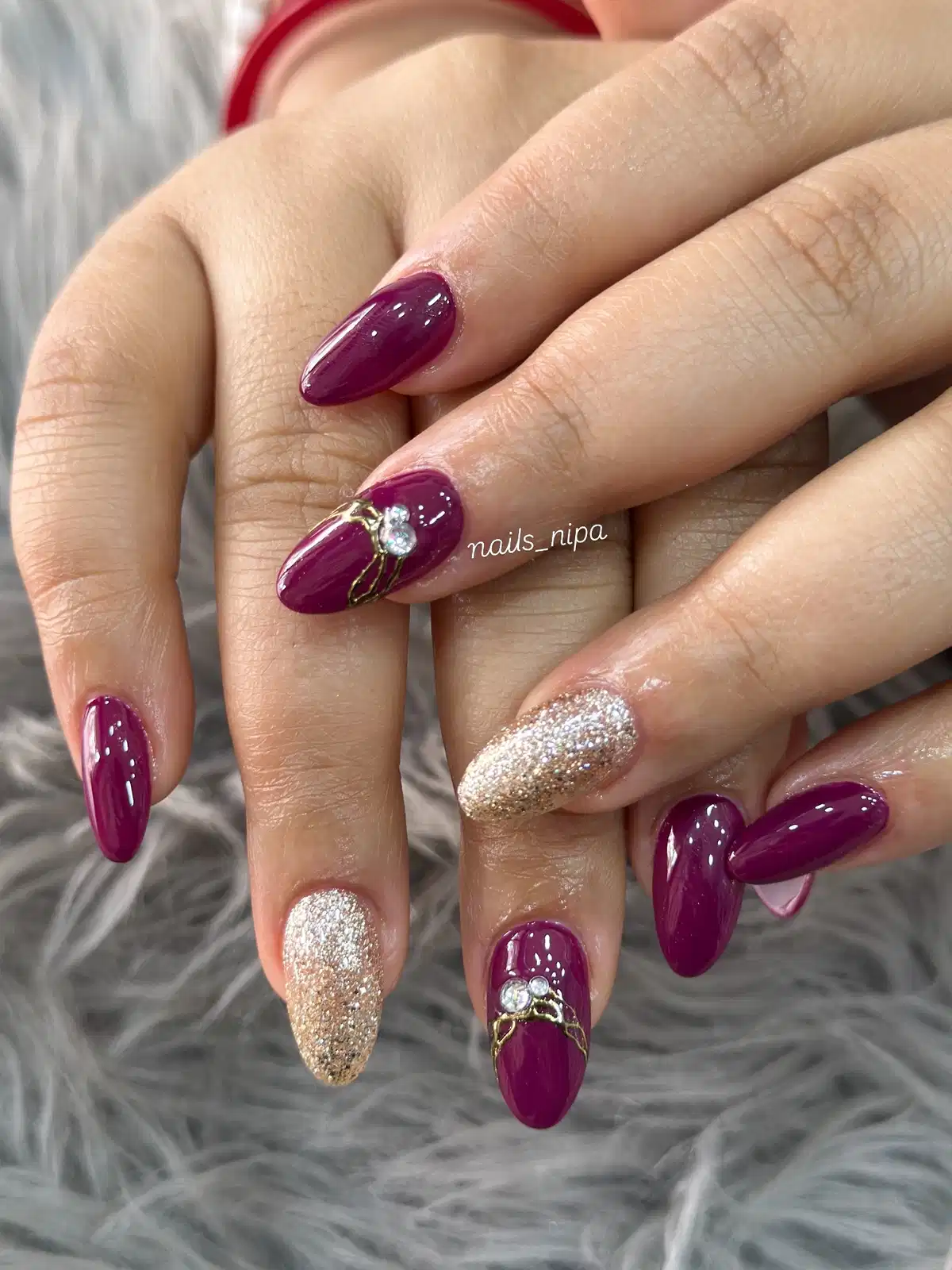 Plum-maroon nails with gold glitter accents and rhinestone details