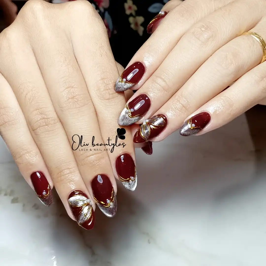 Maroon nails with silver glitter tips, gold curves, and pearl accents