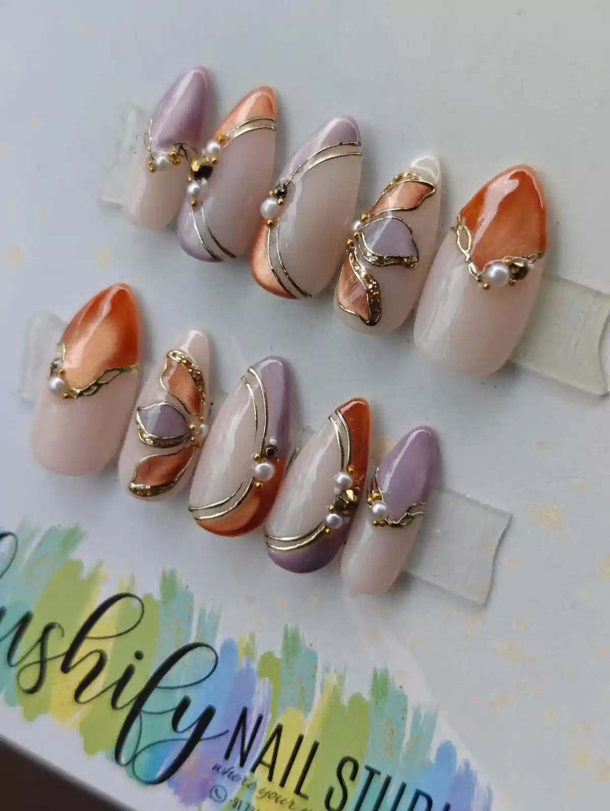 Nude, lavender, and orange-brown nails with gold curves and pearl accents