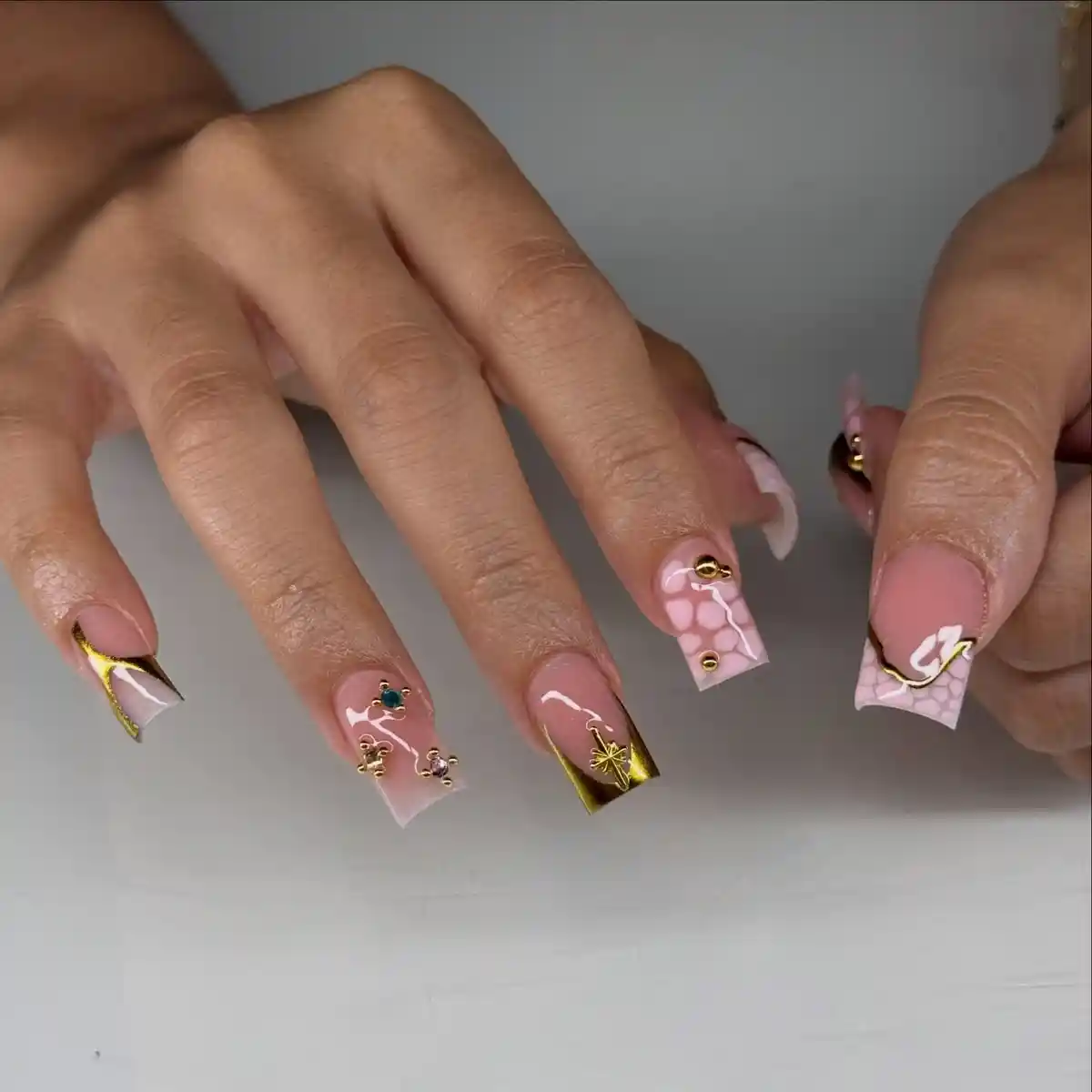 Square pink nails with gold tips, charms, and white pattern accents