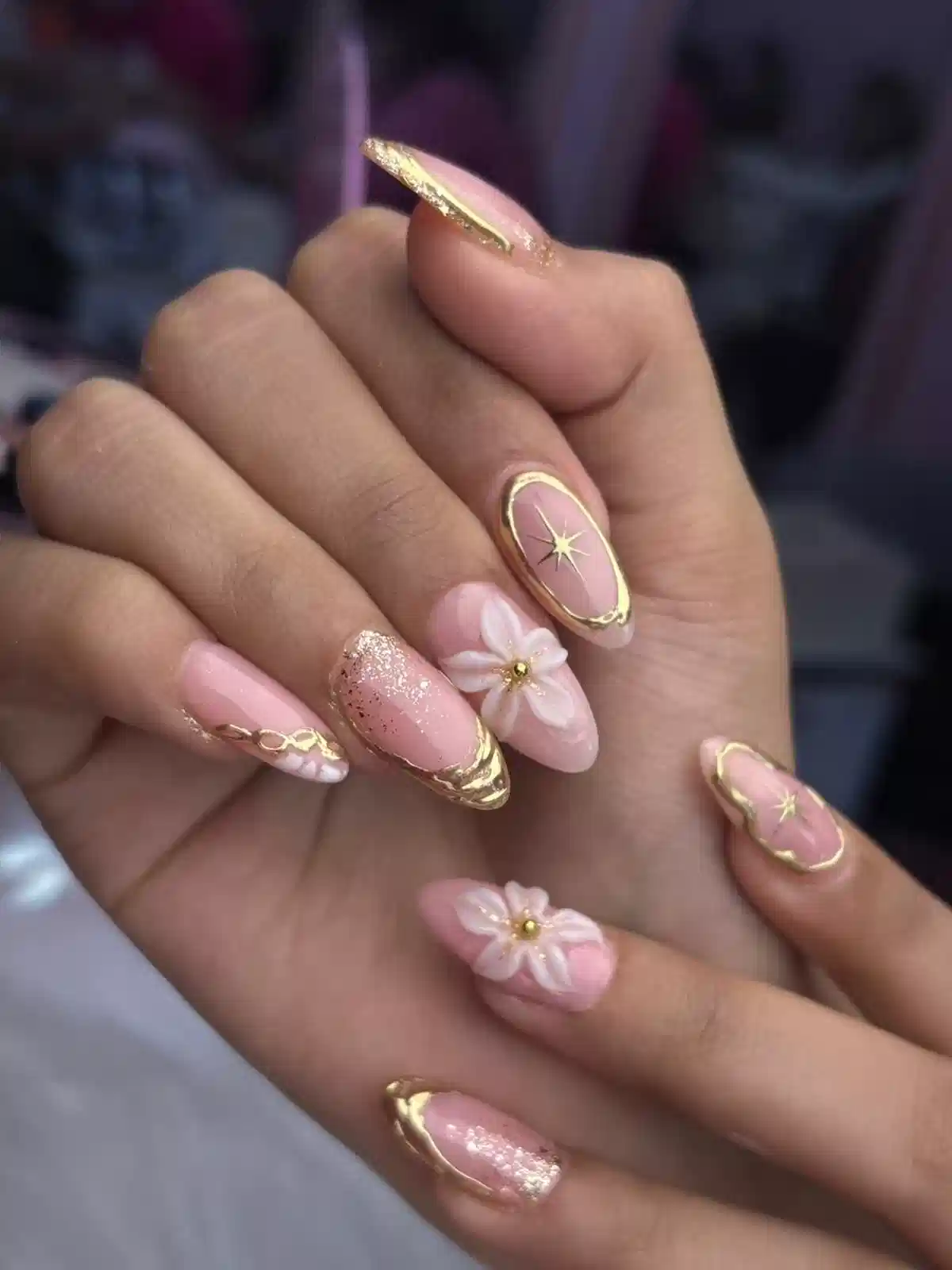 Pale pink almond nails with gold frames, starbursts, and 3D flowers